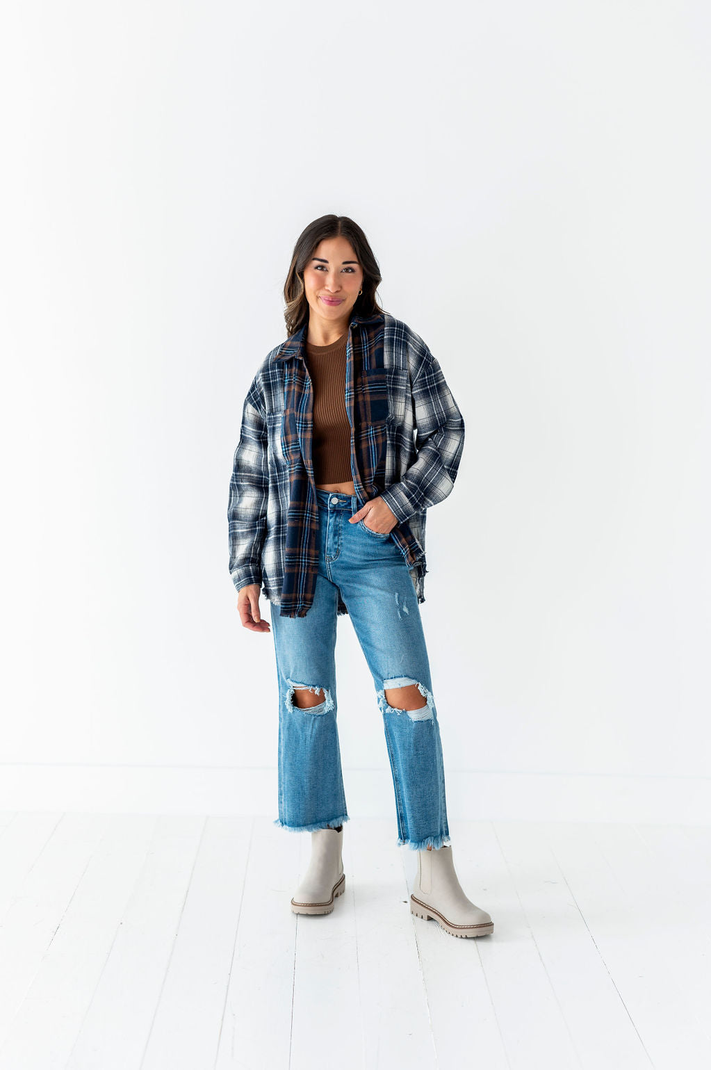 A smiling woman standing in a bright white studio wearing a brown crop top, blue and brown plaid flannel shirt, distressed wide-leg jeans with ripped knees, and light grey lug-sole ankle boots