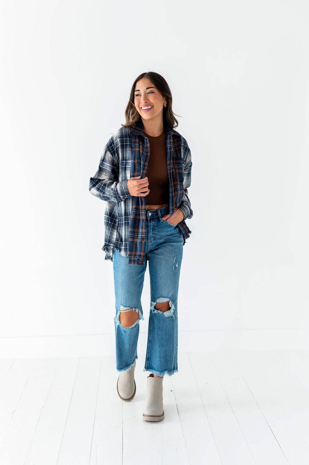 A smiling young woman in an open blue and brown plaid flannel shirt, brown crop top, ripped light blue jeans with frayed hems, and light grey ankle boots stands in a clean white studio