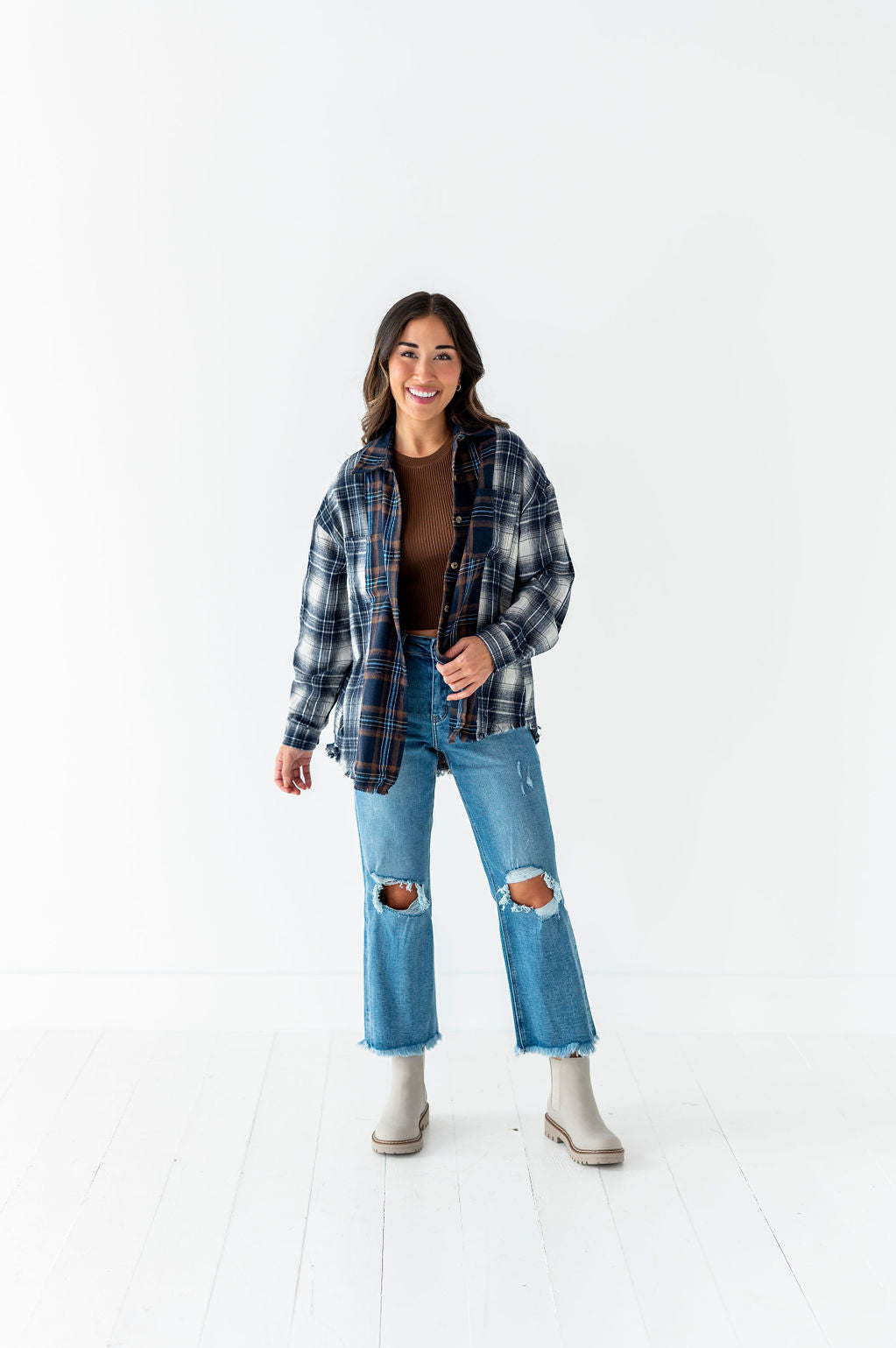Photography, Artist unknown, a smiling woman in a bright white studio wearing an oversized blue and brown plaid flannel shirt, distressed wide-leg jeans with knee rips, and light grey lug-sole Chelsea boots