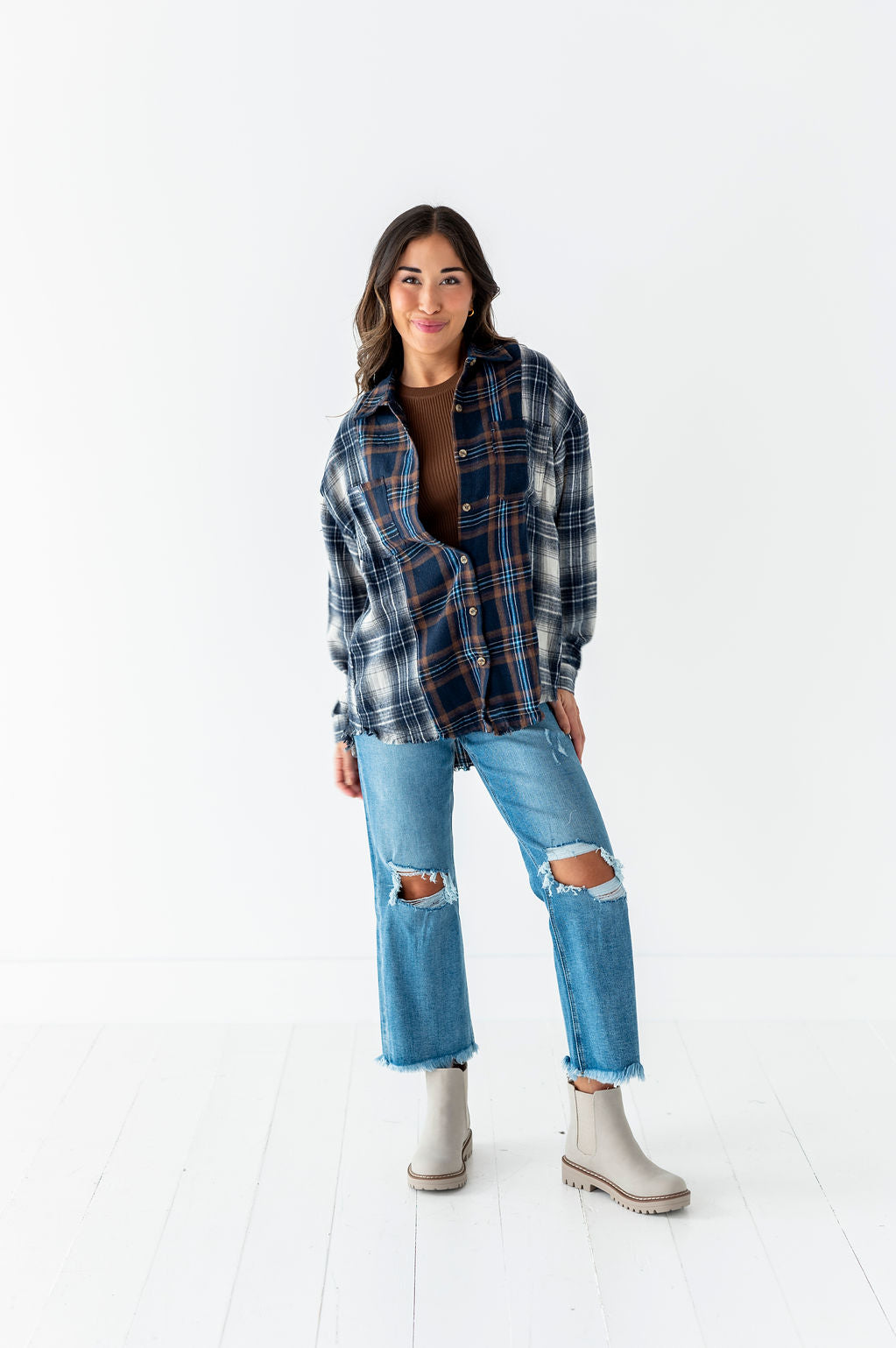 Photograph of a smiling woman in a white studio wearing an oversized dark blue and brown plaid flannel shirt over a brown top light wash ripped jeans with frayed hems and light taupe Chelsea boots