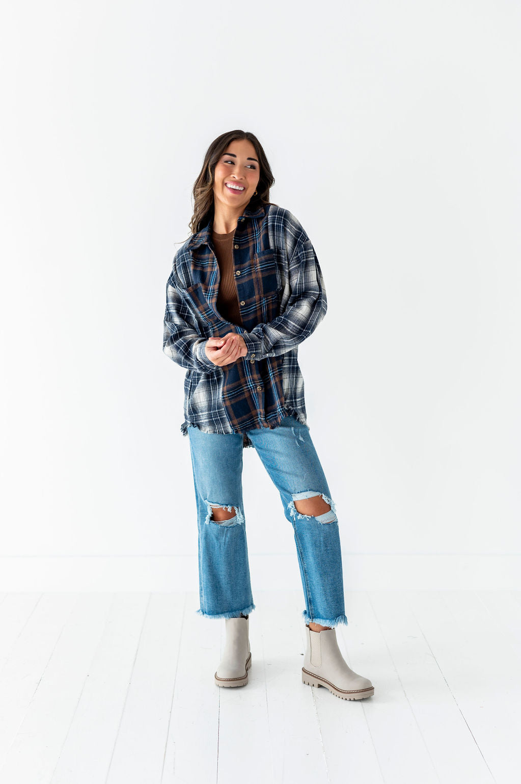 Photograph of a smiling woman in a white studio wearing a dark blue and brown distressed plaid flannel shirt over a brown top, light wash ripped jeans, and light gray lug sole boots