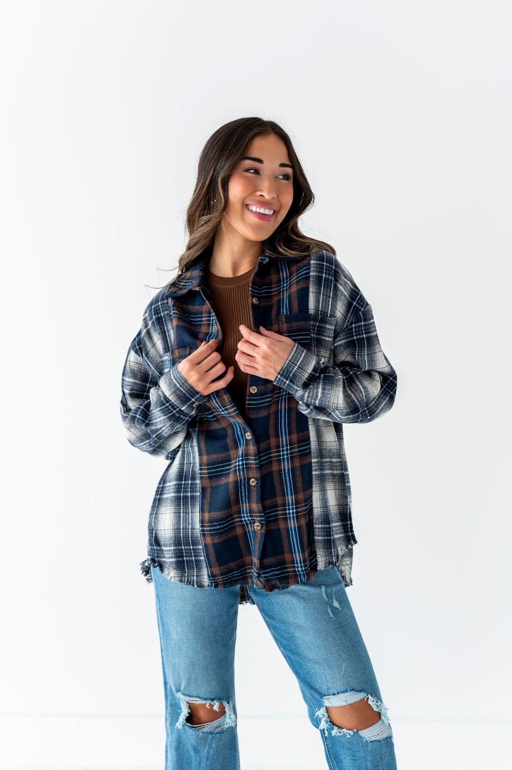 Photograph of a smiling woman in a studio wearing an oversized multi-plaid flannel shirt in blue white and brown over a brown top with distressed light-wash ripped jeans