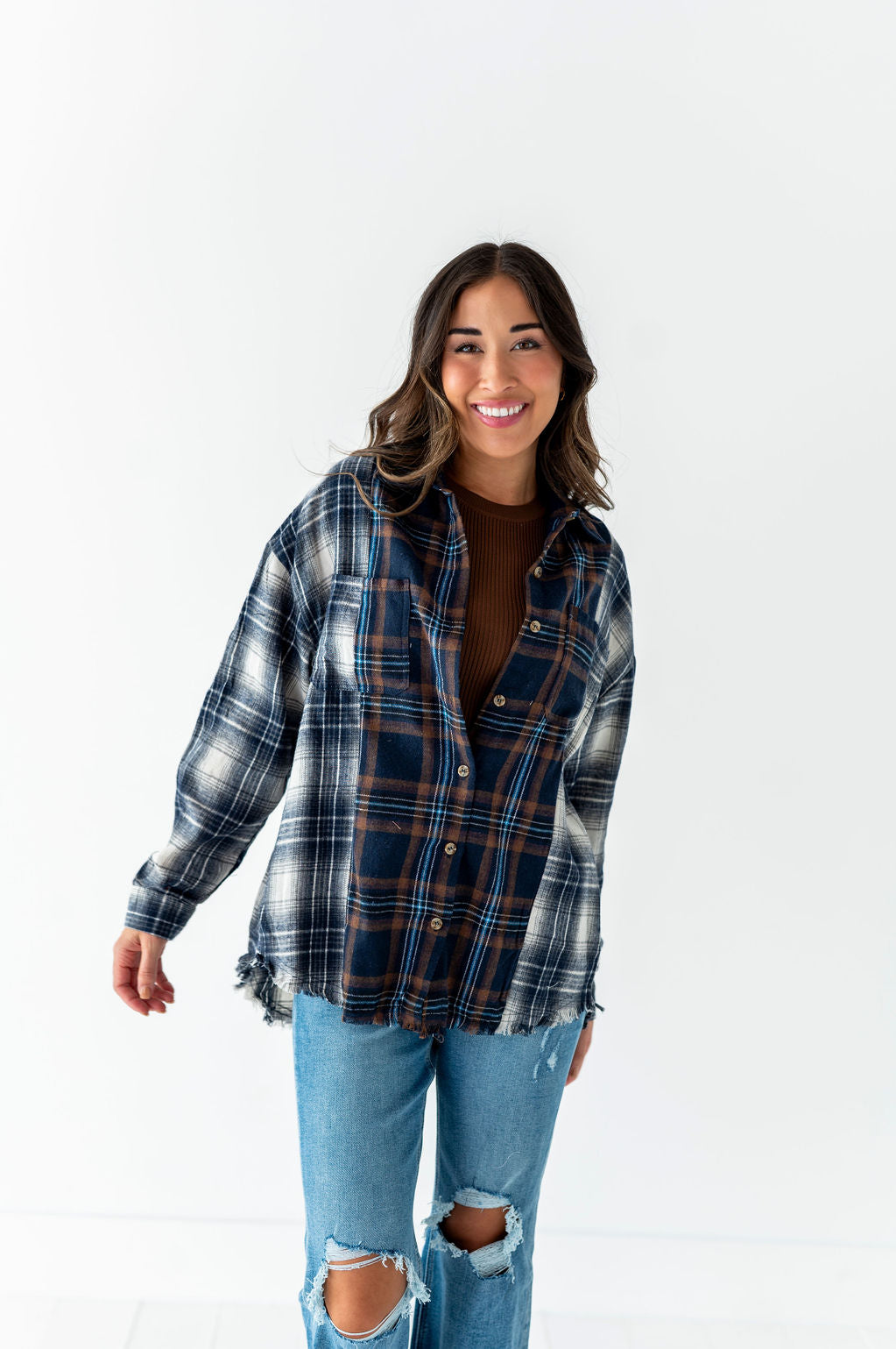 Studio photograph of a smiling woman in an oversized navy blue, white, and brown plaid flannel shirt over a brown top and light wash ripped jeans with knee holes