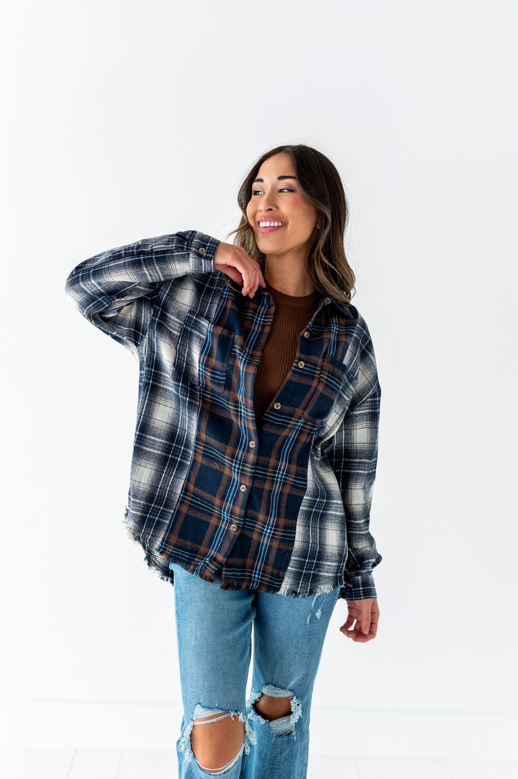 Digital photograph by unidentified artist of a smiling woman in a studio wearing an oversized multi-patterned blue and brown plaid flannel shirt with frayed edges over a brown ribbed top and light wash ripped jeans
