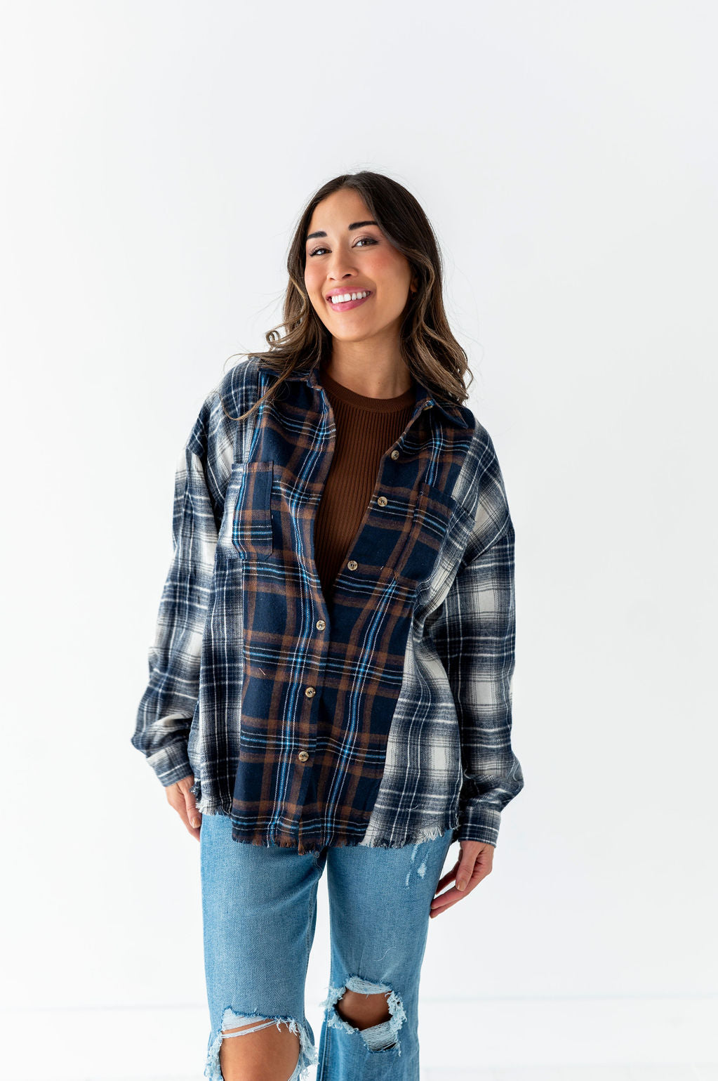 Smiling woman with long dark hair wearing an oversized navy and brown plaid flannel shirt over a brown top and distressed light wash jeans with rips at the knees on a white studio background