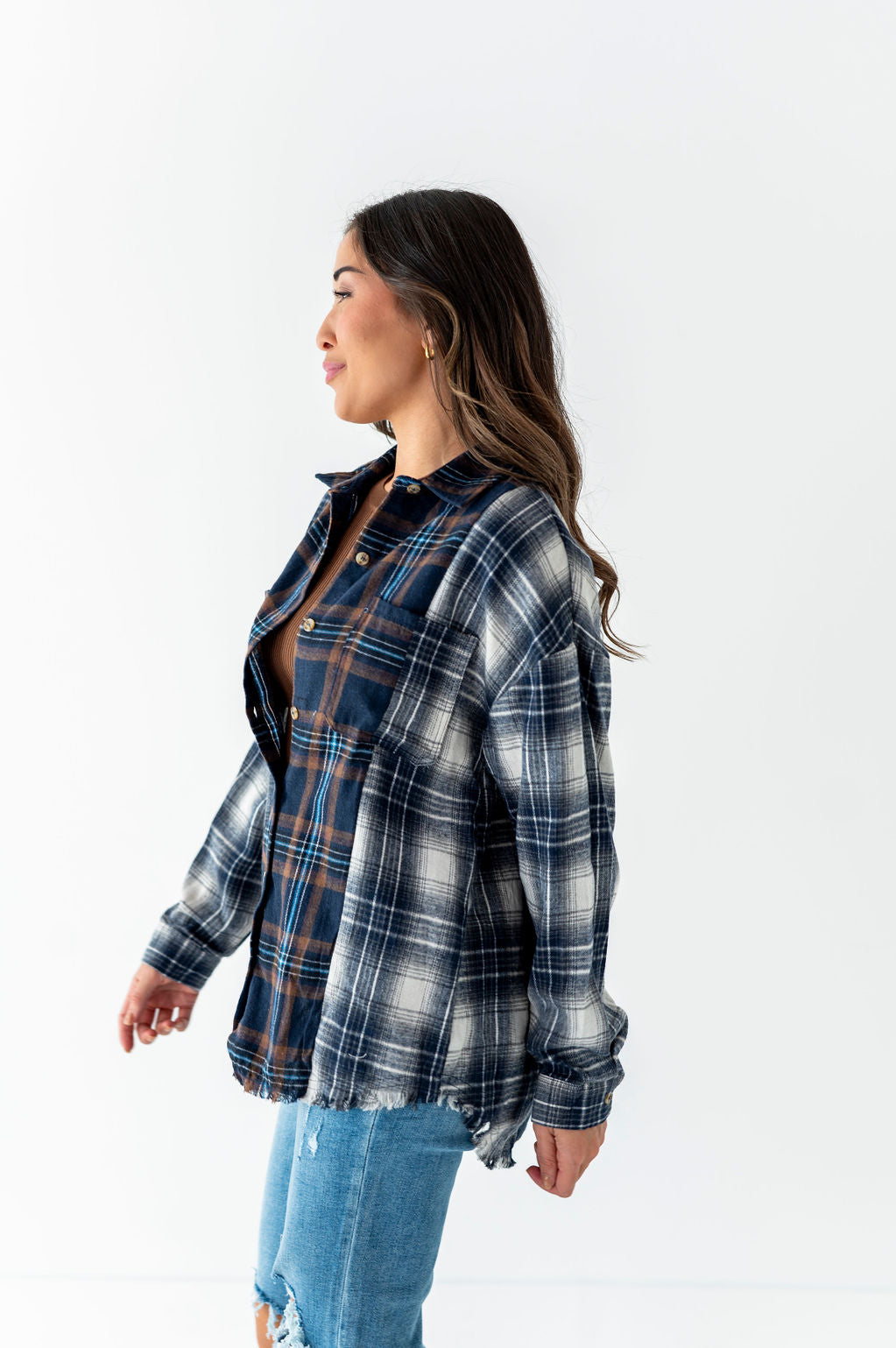A woman wearing an oversized blue white and brown mixed-plaid flannel shirt with a frayed hem over a brown undershirt and light wash ripped jeans against a white background