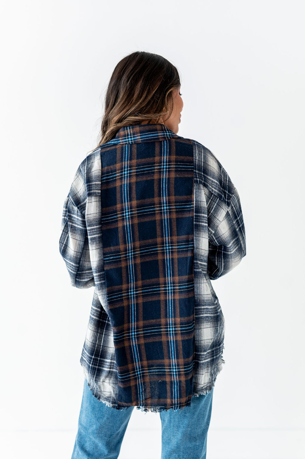 Product photograph of a model from behind against a white background showcasing an oversized color-block flannel shirt with dark blue, brown, and light blue plaid on the back panel contrasting with lighter blue and white plaid on the sleeves, paired with light wash jeans and a distressed bottom hem