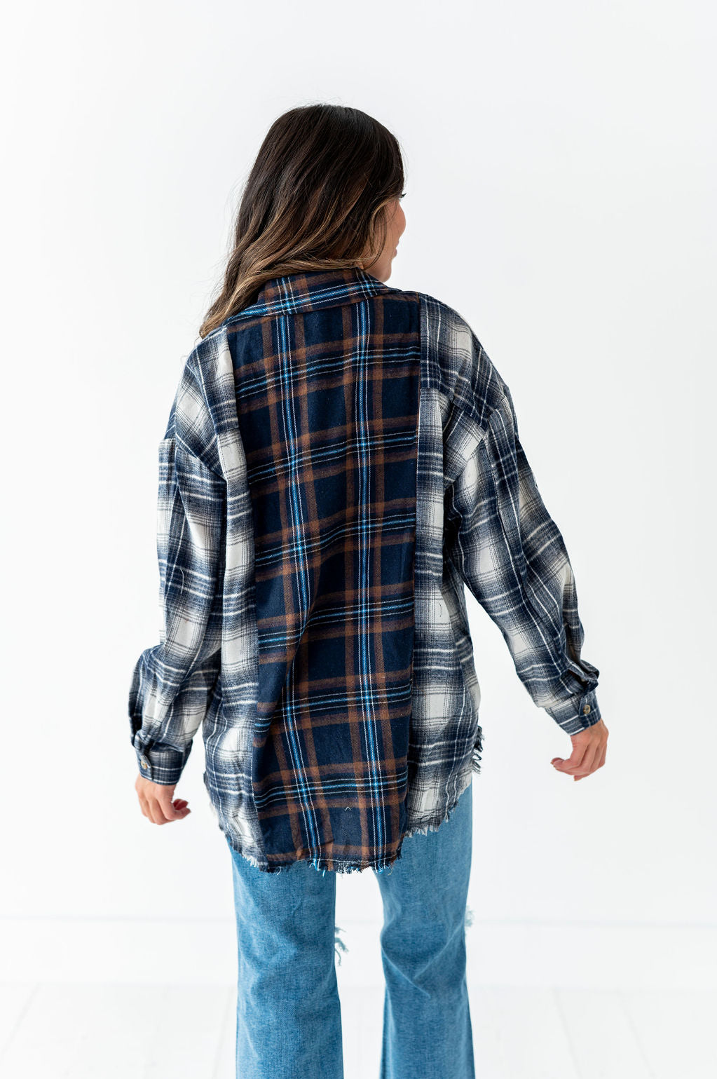 A person viewed from the back wears an oversized navy, brown, and white plaid flannel shirt with a frayed hem and light blue distressed jeans against a clean white studio background