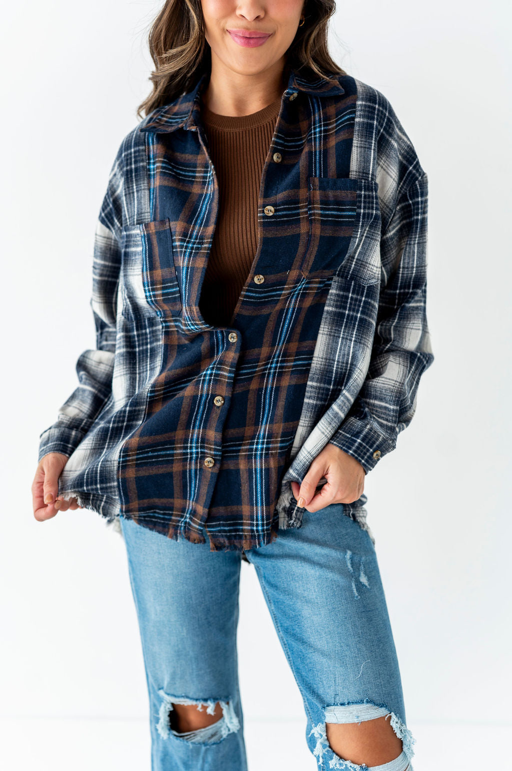 Photograph of a smiling woman in a studio wearing a patchwork navy, brown, and white distressed flannel shirt over a ribbed brown top and light wash ripped jeans