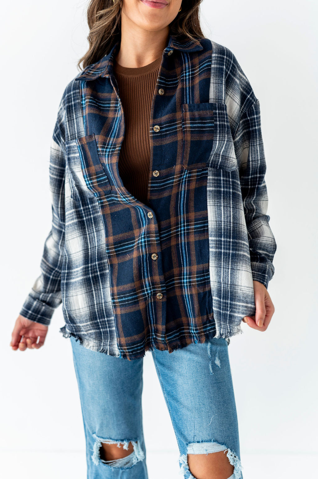 Photograph of a woman in a studio wearing an oversized partially unbuttoned flannel shirt with a frayed hem displaying mixed navy brown and white plaid patterns with light blue accents over a ribbed brown top and distressed light wash jeans