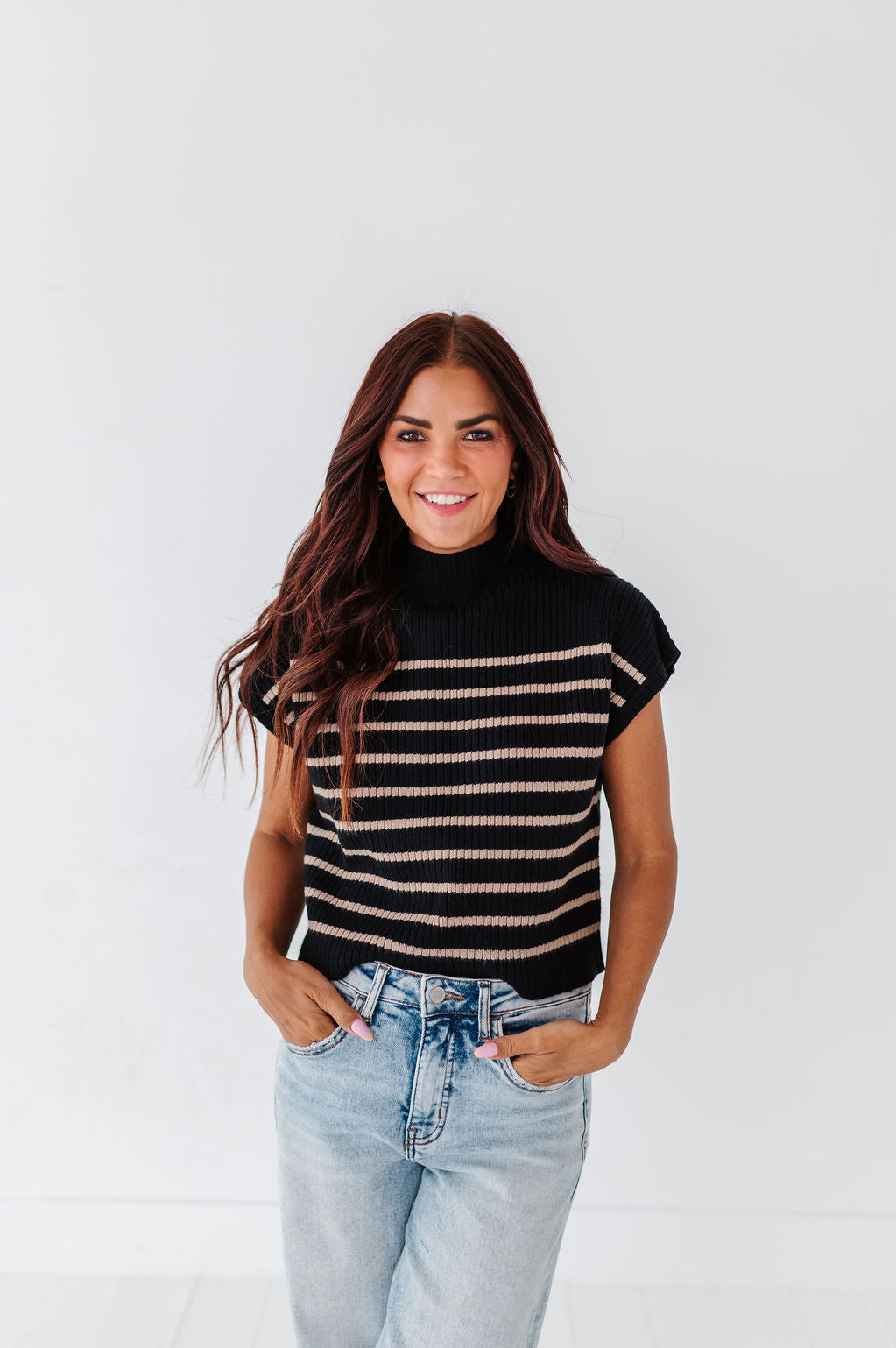Photograph of a smiling woman with long brown hair in a studio wearing a black and beige striped mock-neck sweater, light wash blue jeans with hands in pockets, and pink nail polish