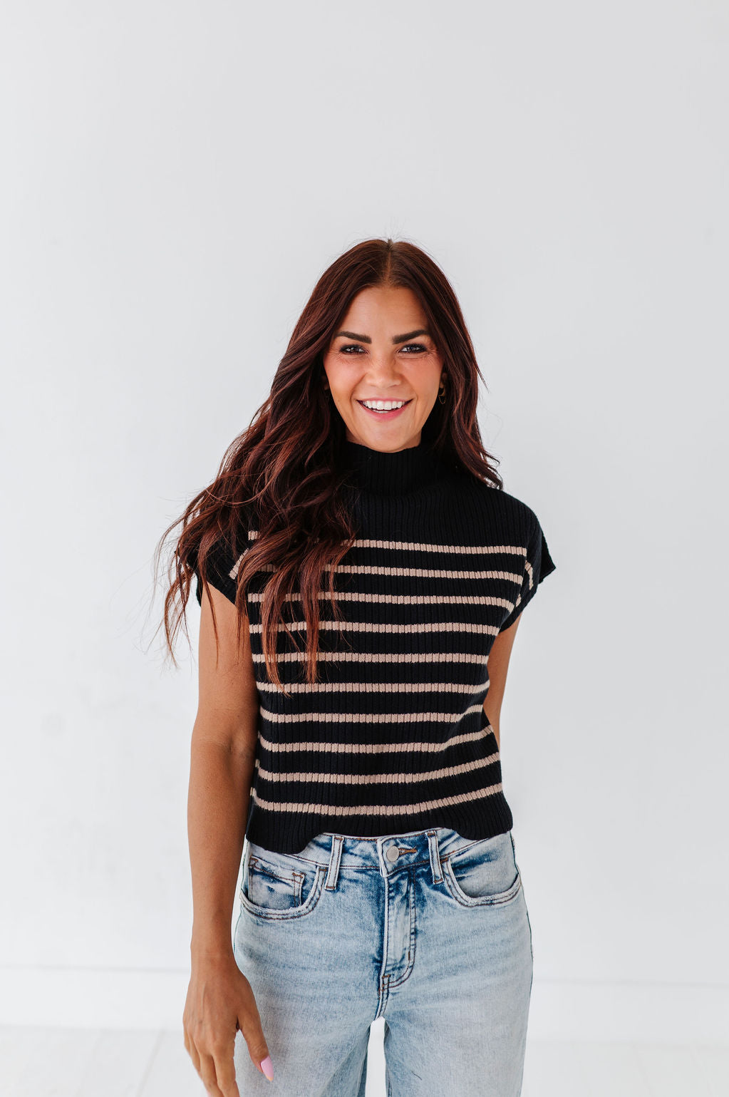A smiling woman with long wavy brown hair wearing a black and tan striped mock-neck sleeveless sweater and light wash blue jeans against a plain white background