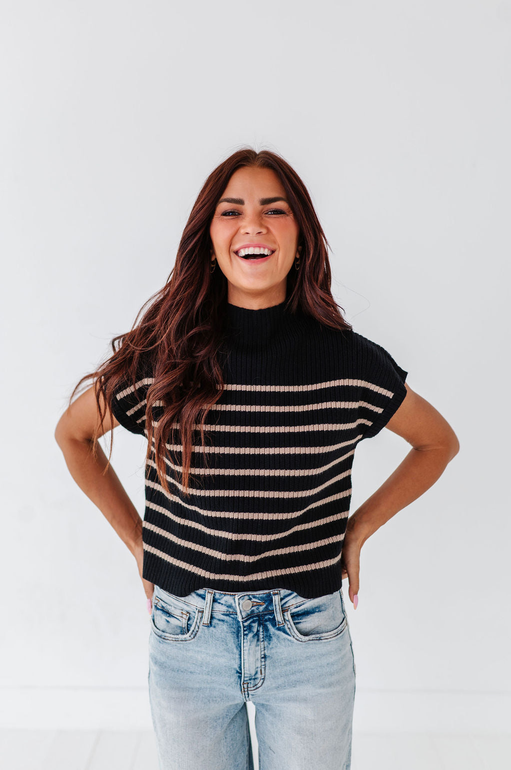 Photograph of a vibrant smiling woman with long brown hair, wearing a black and beige striped mock-neck sweater, light wash jeans, and bright pink eyeshadow against a plain white studio background
