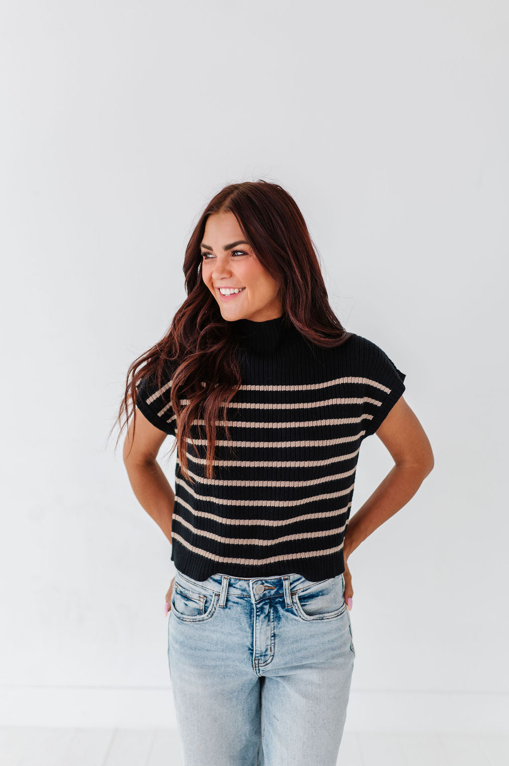 Photograph of a smiling woman with long wavy dark brown hair wearing a black mock-neck sweater with horizontal beige stripes and light wash blue jeans against a plain white studio background