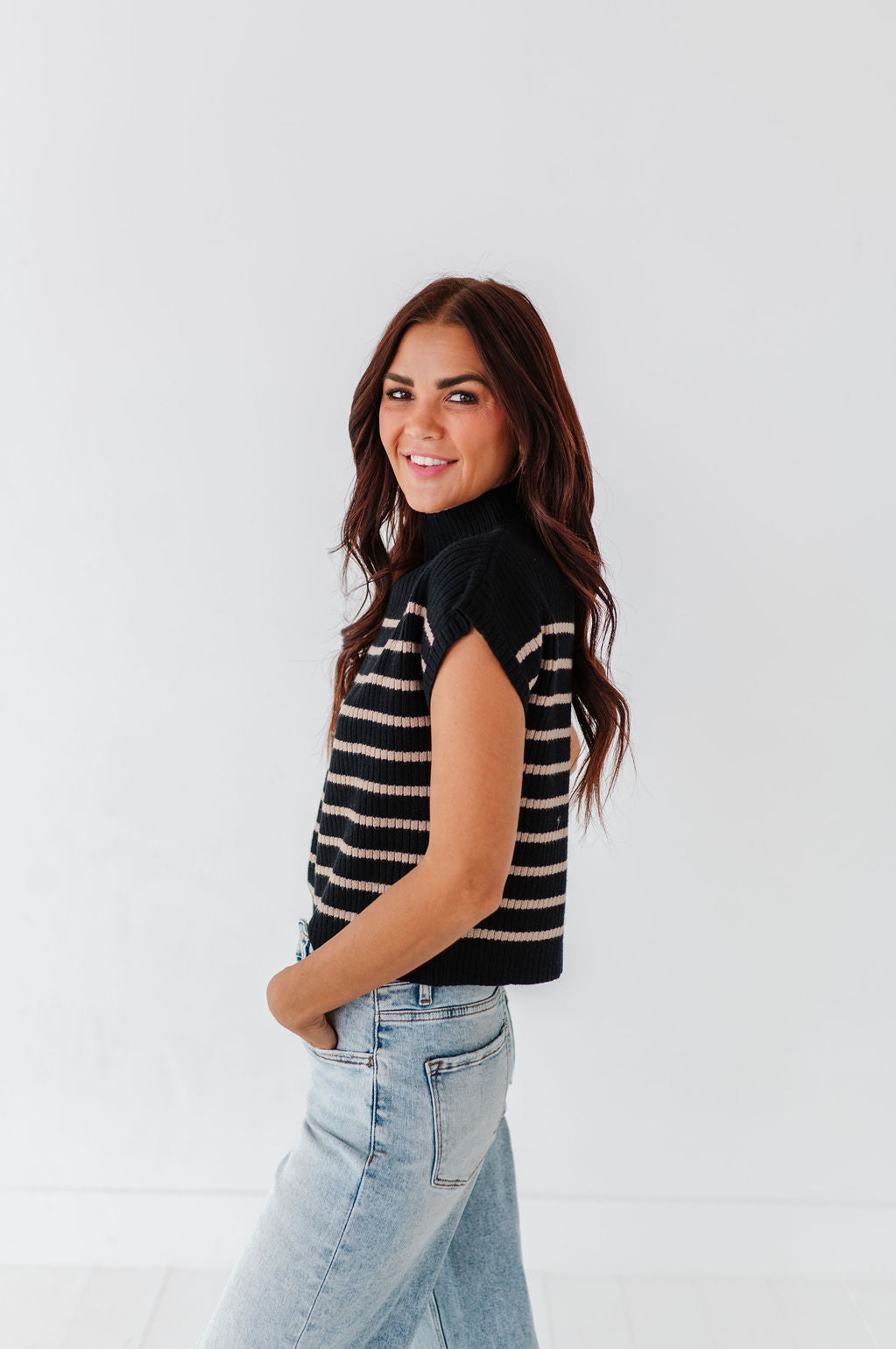 Studio photograph of a smiling woman with flowing dark brown hair wearing a black and beige striped sleeveless mock neck sweater and light wash blue jeans with one hand in her pocket against a crisp white backdrop