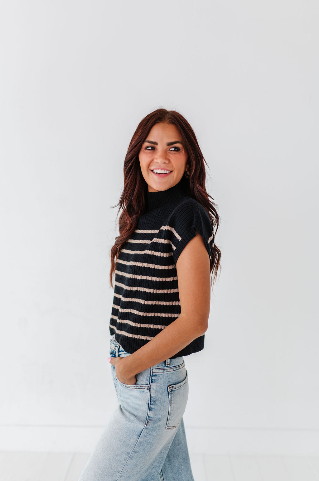 Photograph of a smiling woman with long brown hair wearing a black and beige striped sleeveless turtleneck sweater and light wash jeans with hands in pockets against a white studio background