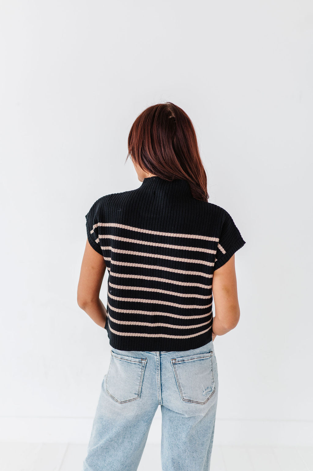 Rear view of a woman with reddish-brown hair wearing a black ribbed short-sleeve turtleneck top with thin tan stripes and light wash distressed jeans against a white studio background