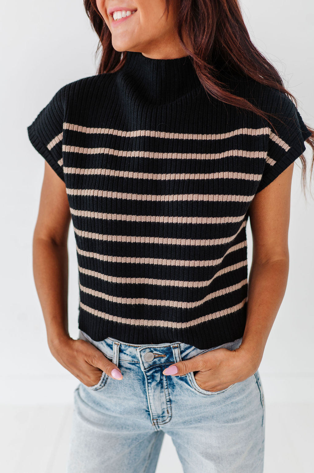 Photograph of a smiling woman in a black ribbed mock-neck sleeveless top with beige stripes and light wash denim jeans, her hands in pockets displaying pink manicured nails