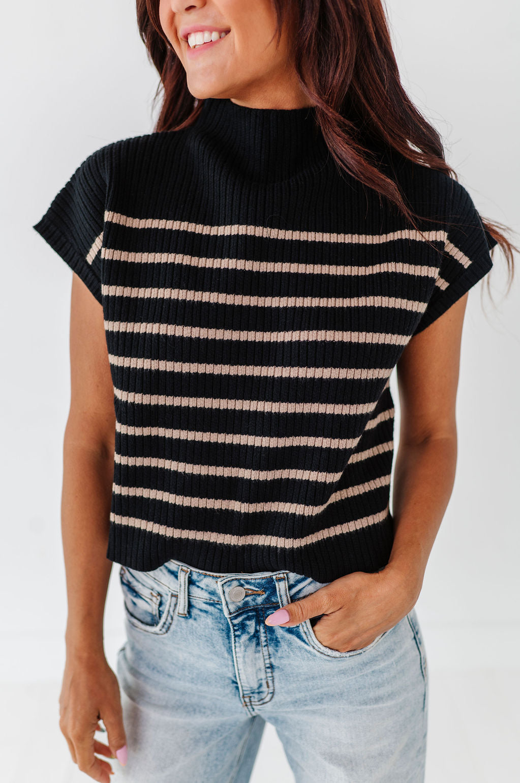 Smiling woman with long dark hair modeling a black high-neck short-sleeved top with thin beige horizontal stripes and light wash blue jeans showcasing pink manicured nails against a clean white background