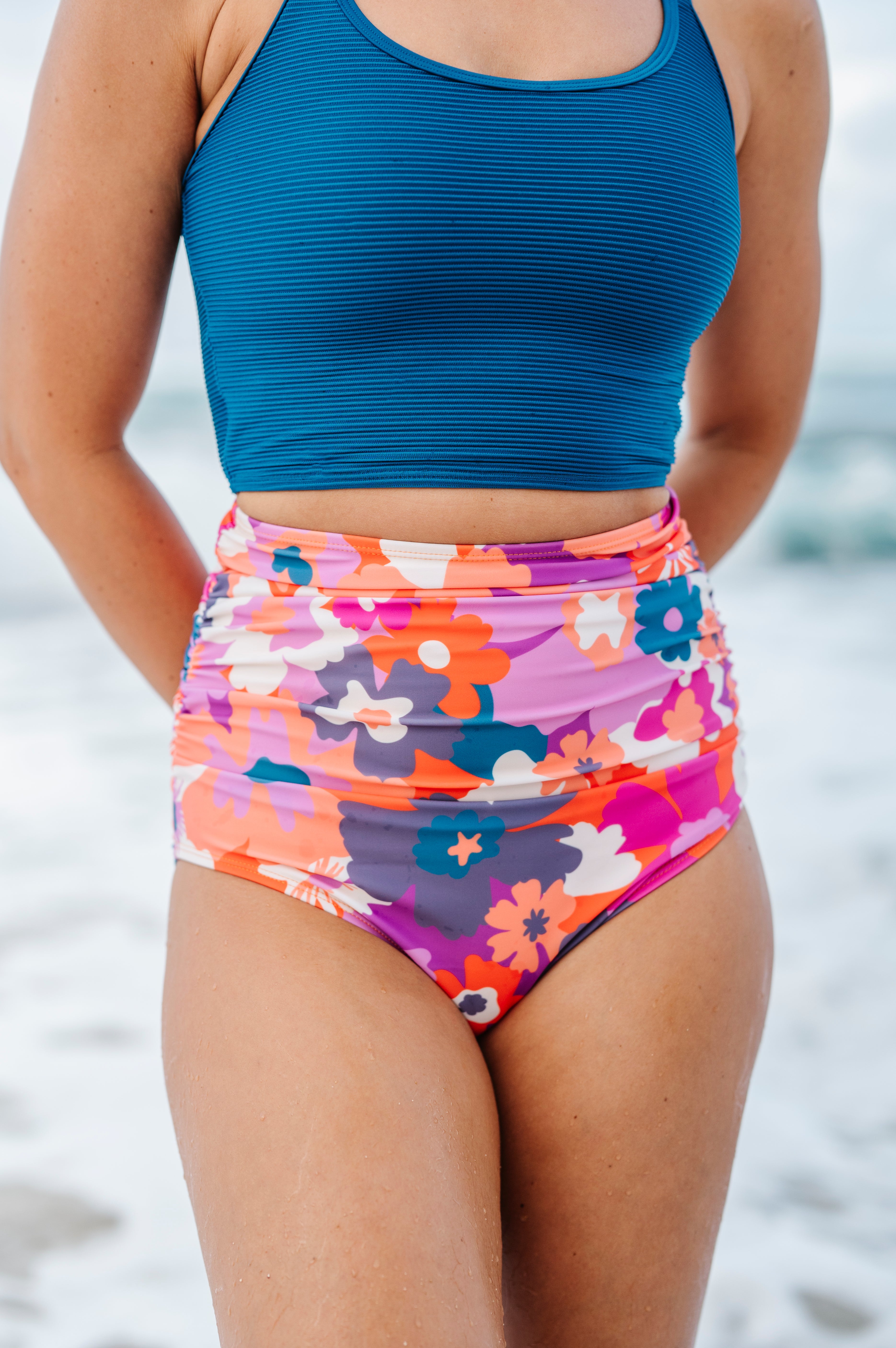 Close-up of a person wearing a ribbed blue crop top and high-waisted swimsuit bottoms with a vibrant pink, orange, purple, and blue floral pattern, standing on a beach with hands behind their back and water droplets on their skin
