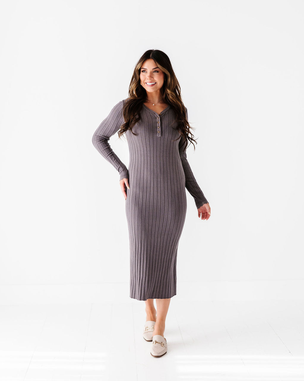 A smiling woman in a long-sleeved ribbed grey midi dress with a Henley neckline and light beige flat mules with gold hardware stands in a bright white studio