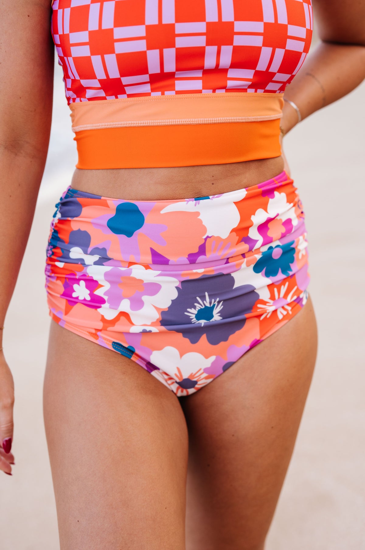 Photograph of a person in a vibrant two-piece swimsuit featuring an orange and lavender checkerboard crop top, a solid orange midriff band, and high-waisted bottoms with a multicolored floral pattern in pink, orange, purple, and blue