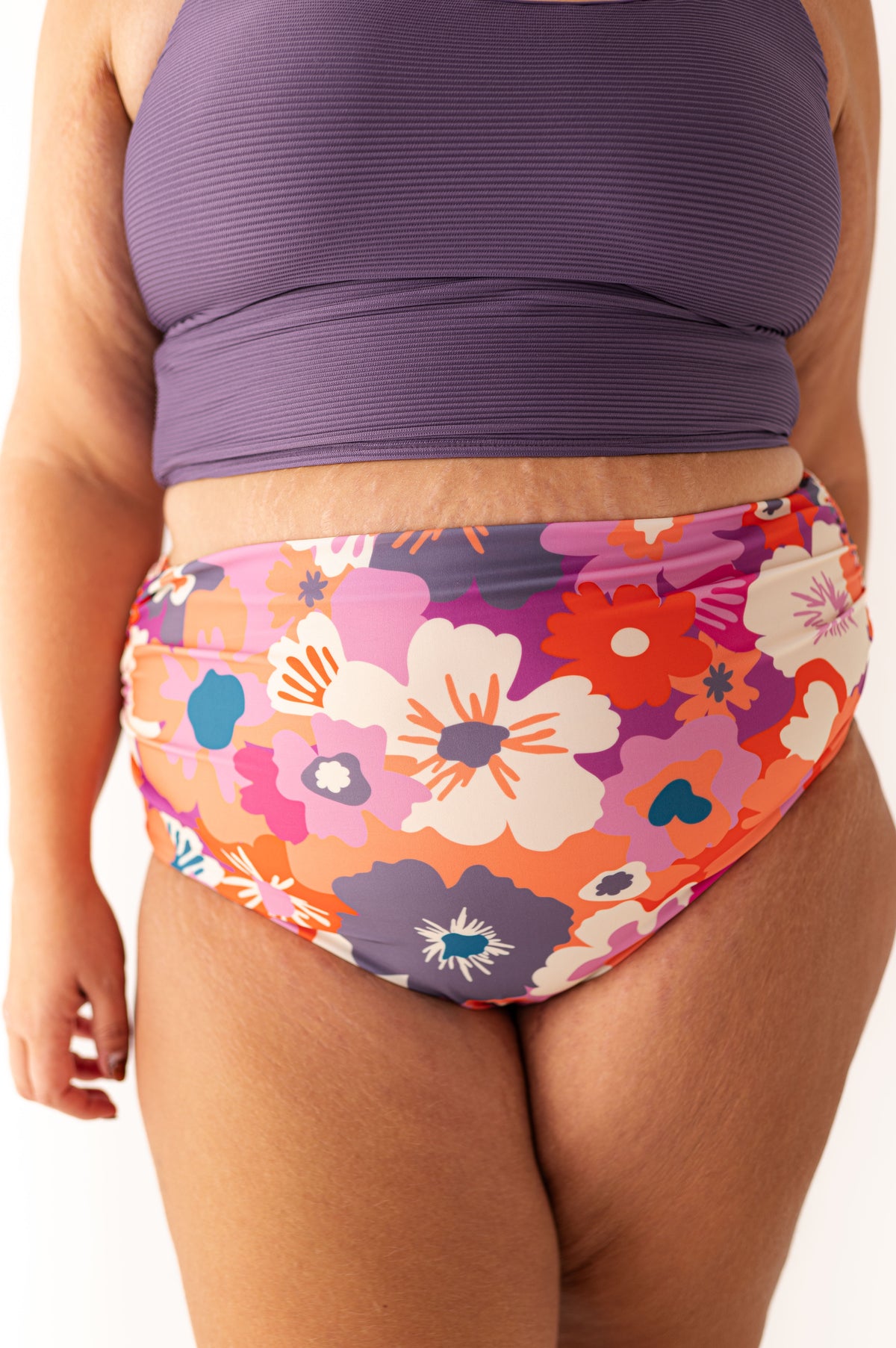 Close-up of a person's midsection wearing a ribbed purple bikini top and a high-waisted swimsuit bottom with a vibrant orange pink purple and white floral pattern against a white background showing natural skin texture
