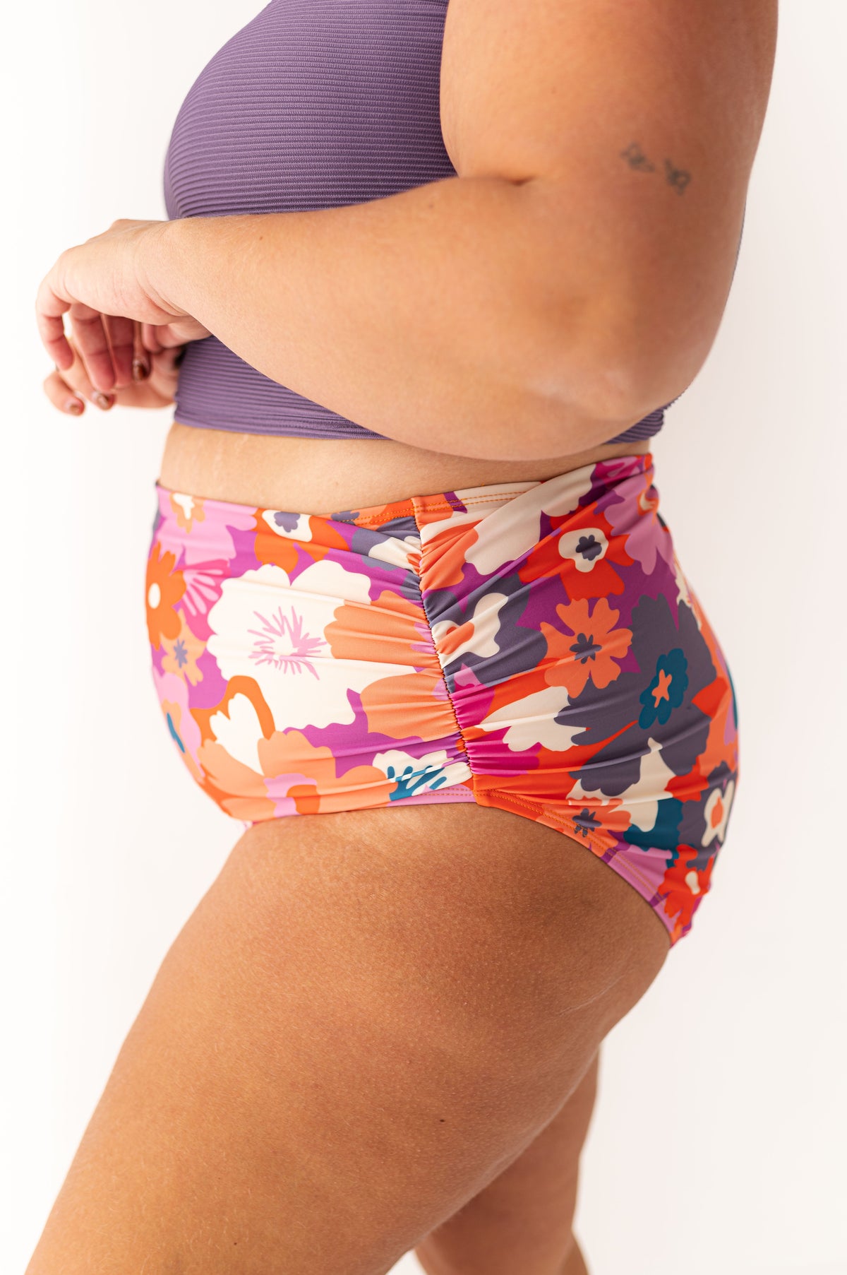 Woman's midsection and upper thigh against a white background, wearing a purple ribbed swim top and a high-waisted swimsuit bottom with a vibrant floral pattern in orange, pink, purple, white, and teal, featuring ruched sides and visible stretch marks