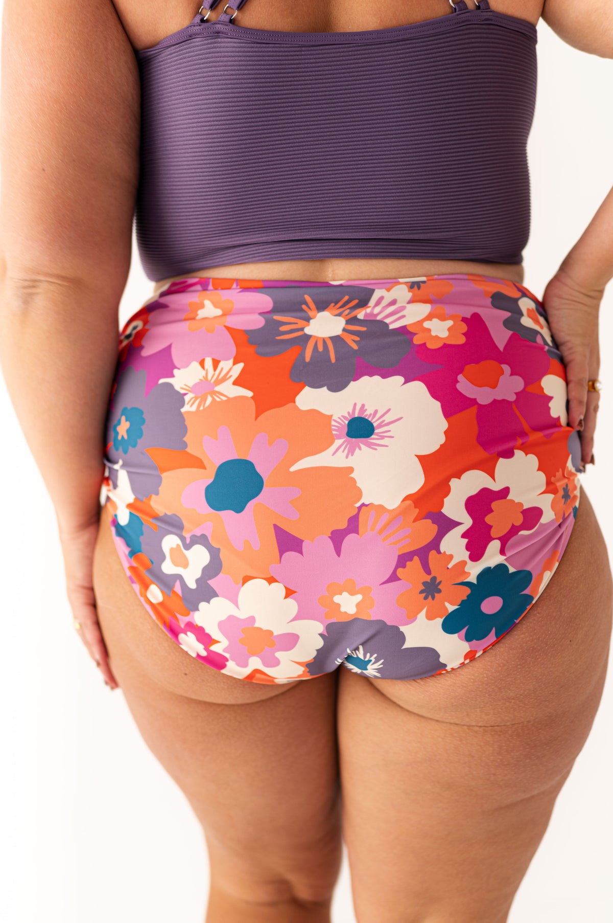 Photograph of the back view of a person wearing a purple ribbed bikini top and high-waisted swim bottoms with a vibrant retro floral pattern in shades of orange, pink, lavender, and teal