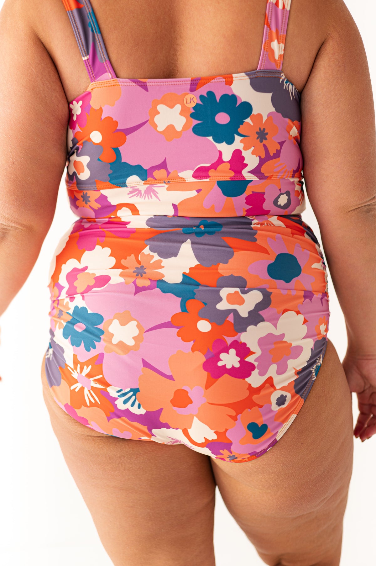 Product photo of a woman's back wearing a vibrant pink orange teal and purple floral print swimsuit with a high-waisted ruched bottom and a subtle LK logo against a white studio background