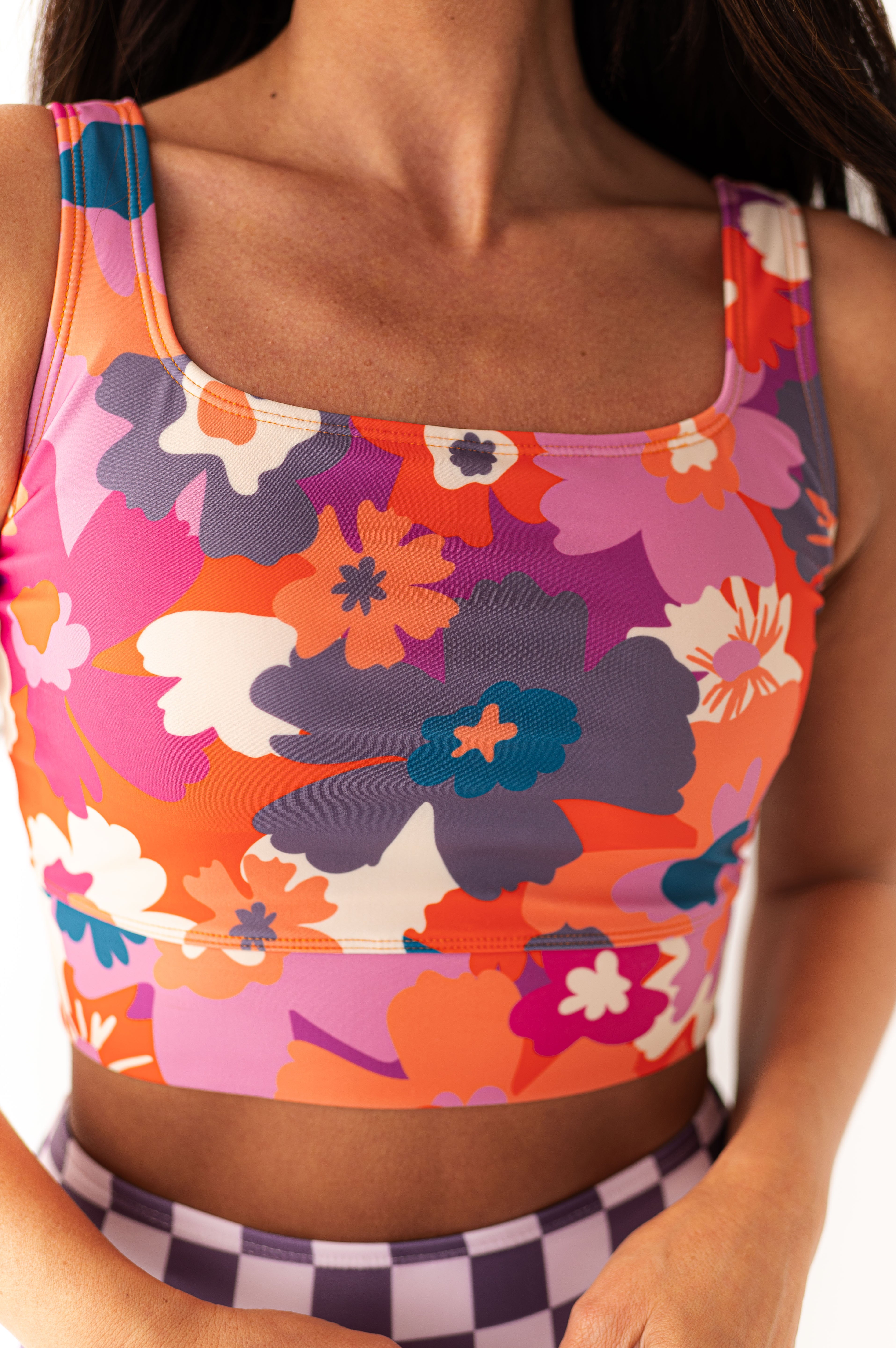A woman modeling a vibrant floral print cropped tank top with orange pink purple and teal flowers paired with a purple and white checkered bottom