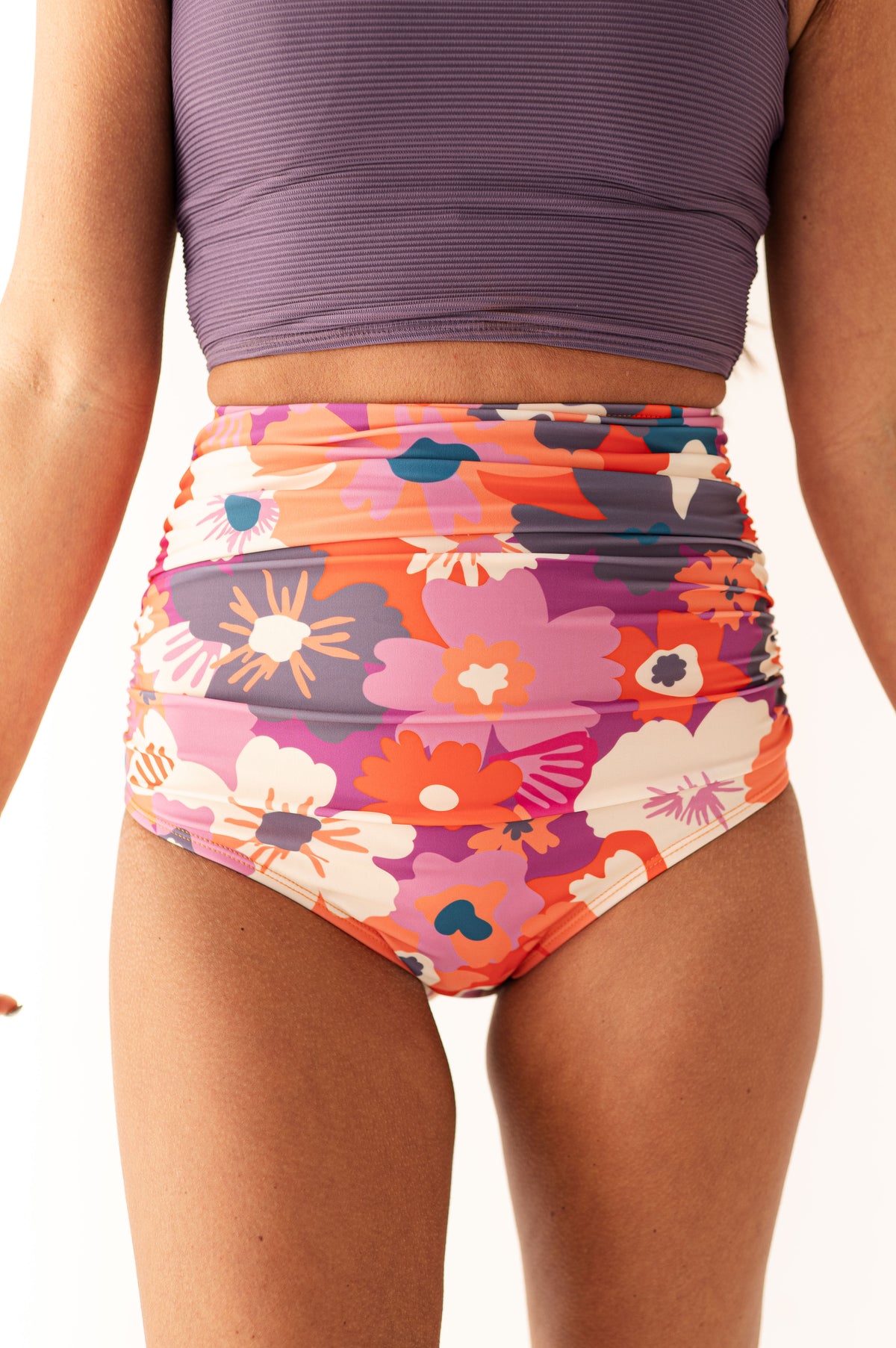 Product photography of a woman in a purple ribbed crop top and high-waisted ruched swimsuit bottom with a vibrant pink orange and purple floral pattern
