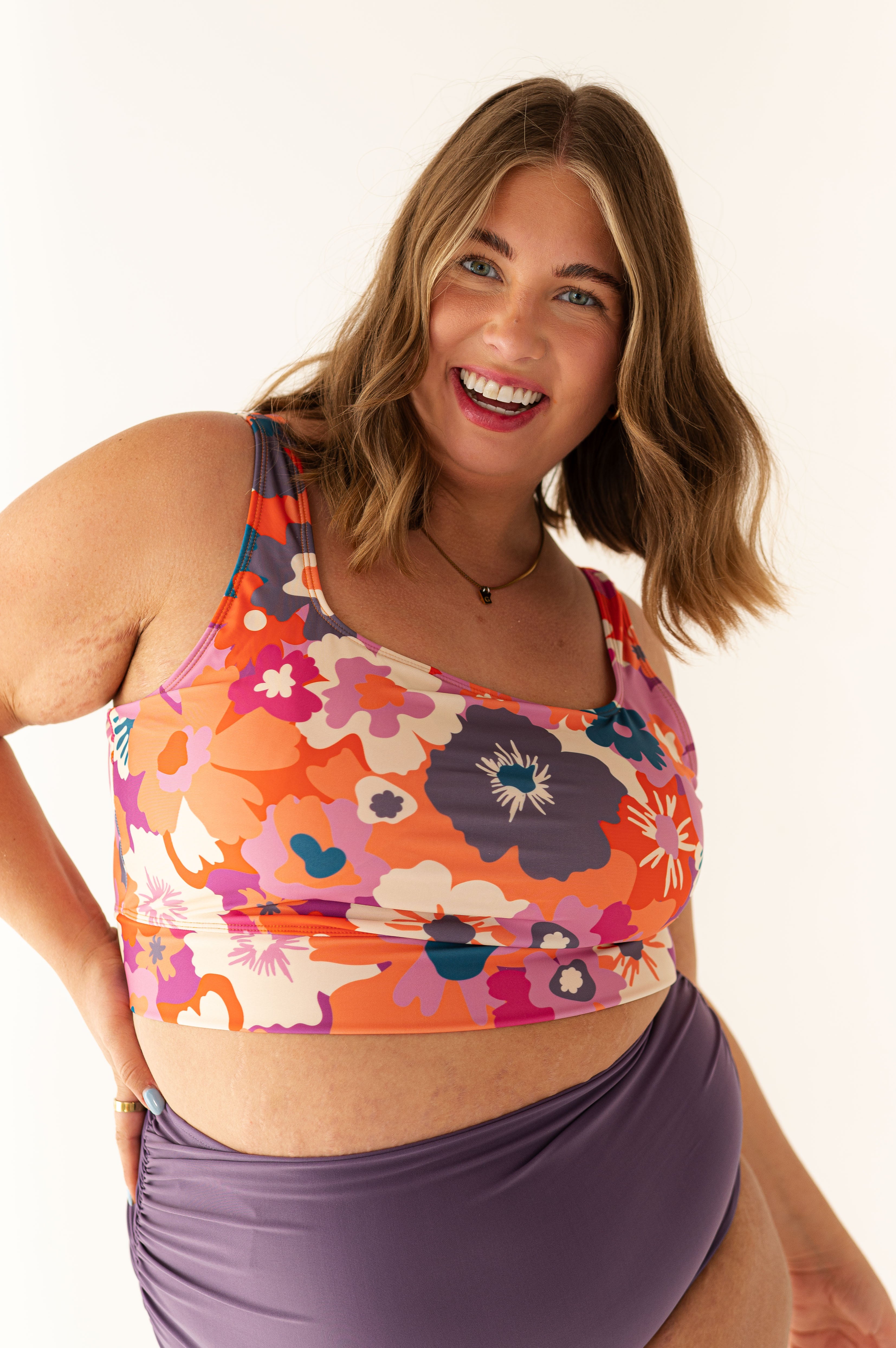 Photograph of a smiling plus-size woman wearing a vibrant orange pink and purple floral crop top and dark purple ruched bikini bottoms with bright blue eyes and a radiant smile against a clean white background