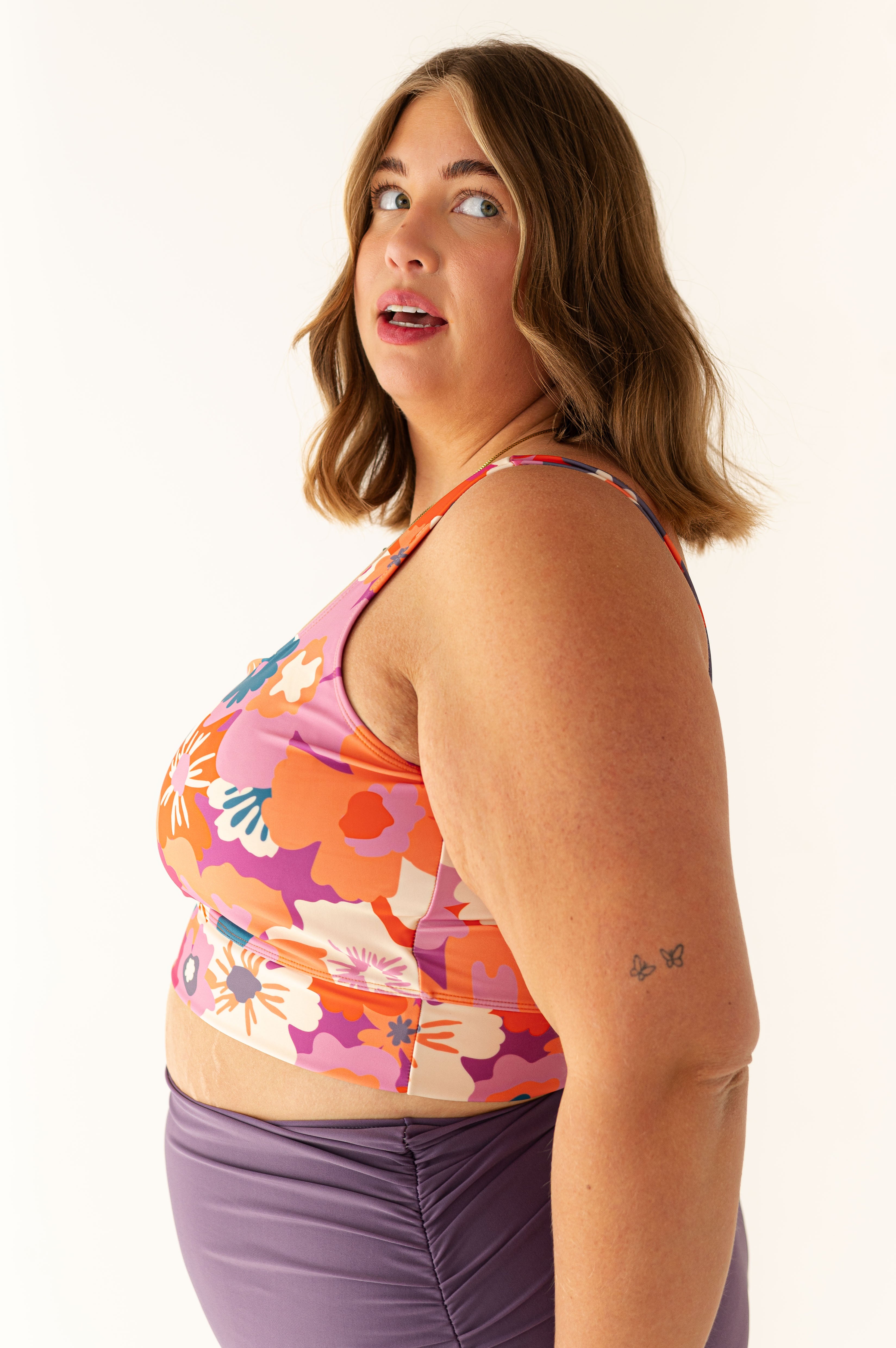 Photograph of a plus-size woman with light green eyes looking up, wearing a vibrant floral print top and ruched purple bottoms, with two small butterfly tattoos on her right arm against a white studio background