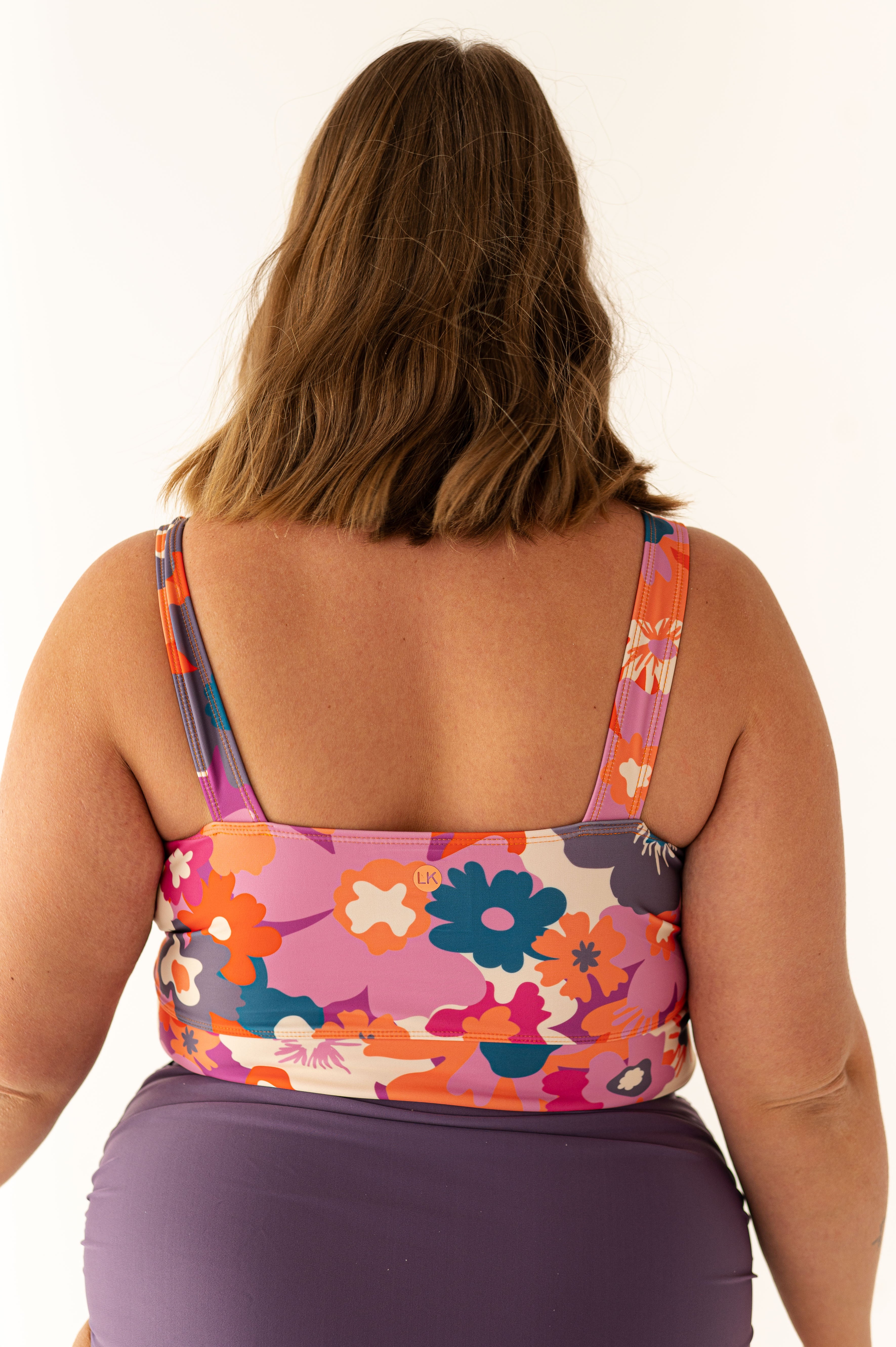 Photograph of a woman from the back against a white studio background, showcasing her brown wavy hair, a vibrant retro floral swimsuit top in purple, orange, teal, and white with a small orange LK logo, and solid dark purple bottoms
